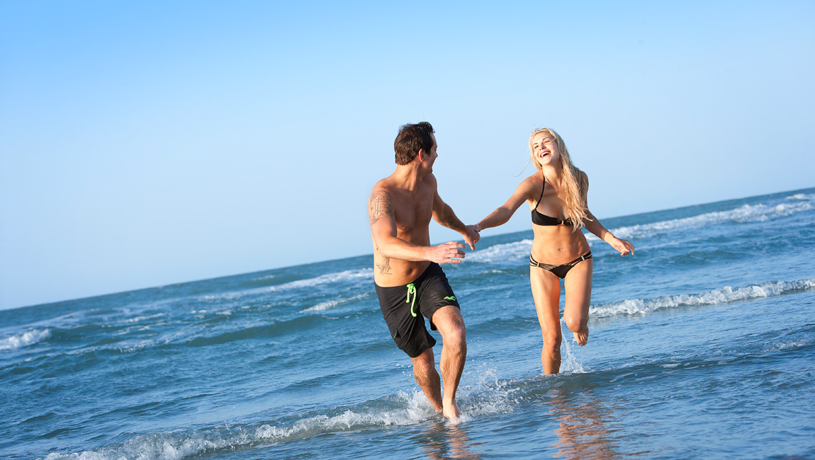 A man and woman run through the water on a beach.