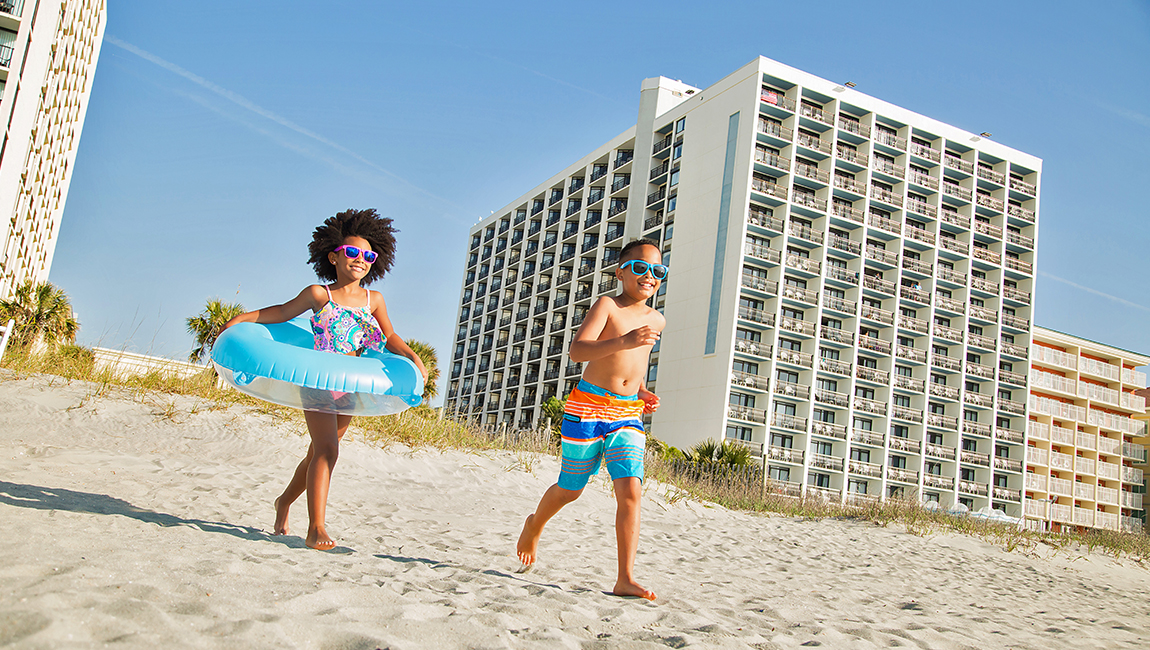 Two children in swimwear running on the beach with a hotel building in the background.