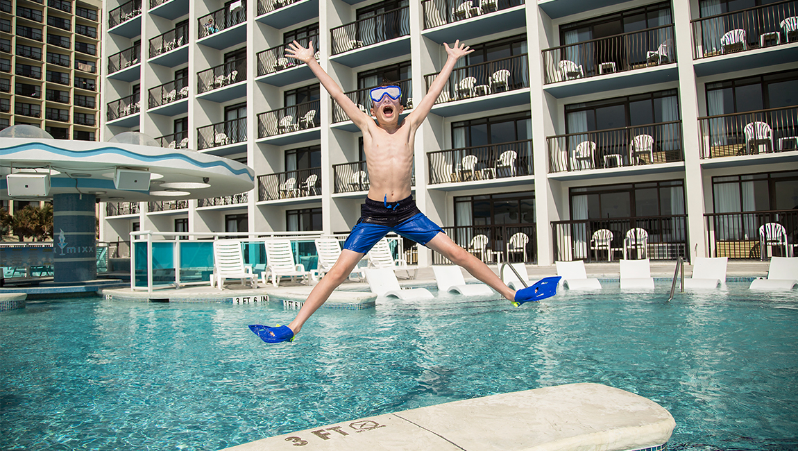 A young boy wearing flippers and goggles jumping into a hotel pool.