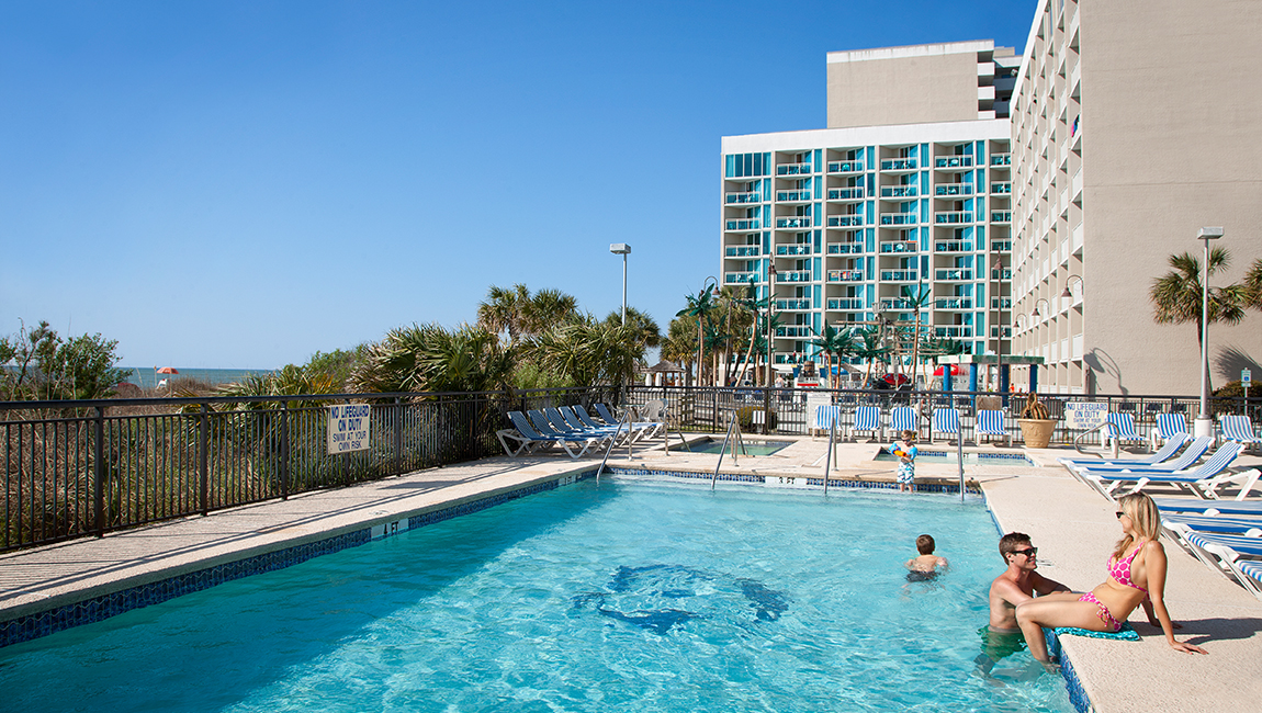 A swimming pool with people in it, surrounded by lounge chairs and buildings.