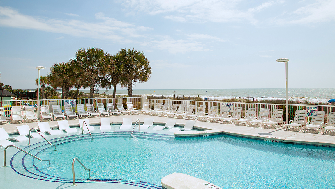 A large pool with white lounge chairs and palm trees in the background, overlooking the ocean.