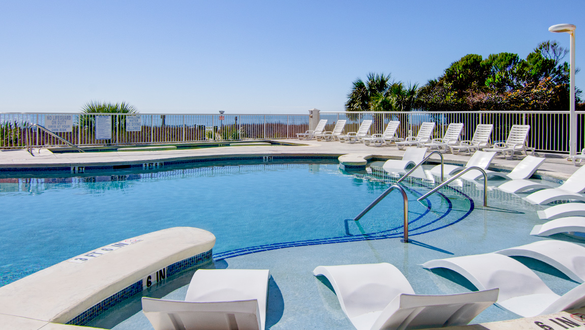 A swimming pool with white lounge chairs and a fence overlooking the ocean.