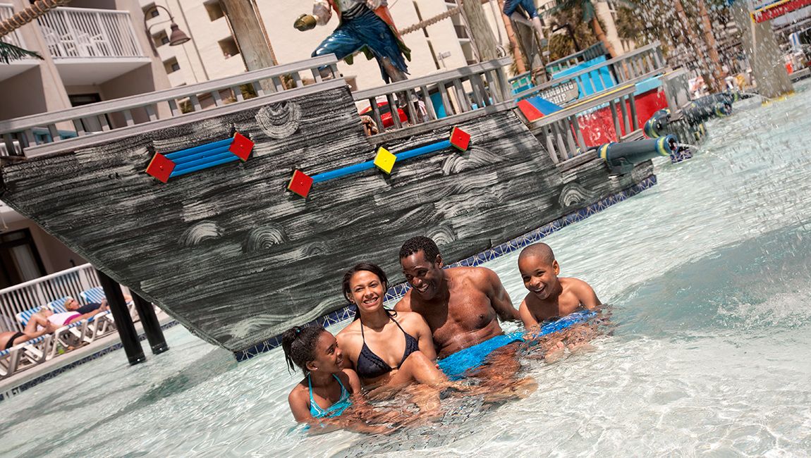 A family of four enjoys a pool with a pirate ship water feature.