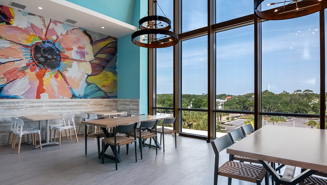 A dining area with tables and chairs, a large mural on the wall, and floor-to-ceiling windows.