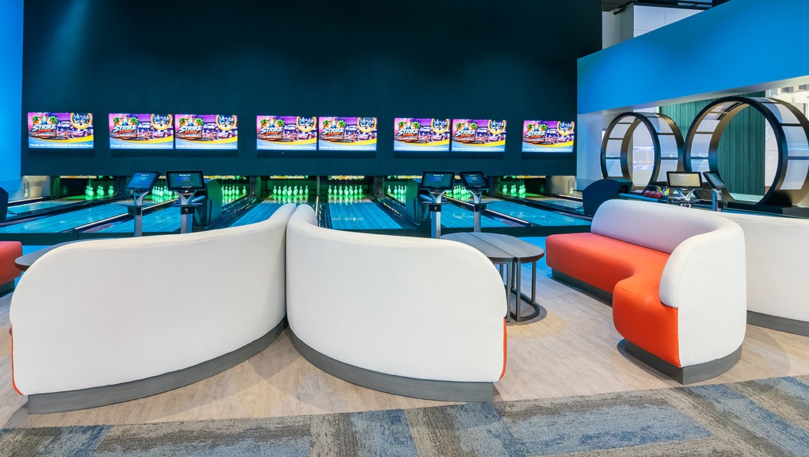 A modern bowling alley with blue lanes, white and orange seating, and video screens on the wall.
